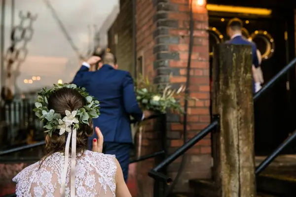 A bride with a floral crown walks up brick steps, following a groom in a blue suit toward a rustic venue entrance. Soft ambient lighting glows.