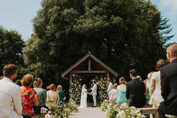 An outdoor wedding ceremony beneath a large tree, featuring a floral-decorated arch and guests seated on rustic benches.