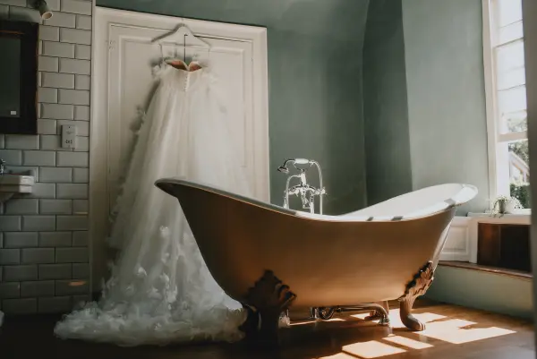 A vintage clawfoot bathtub sits beneath a sunny window, showcasing a stunning wedding dress elegantly hanging on the wall.