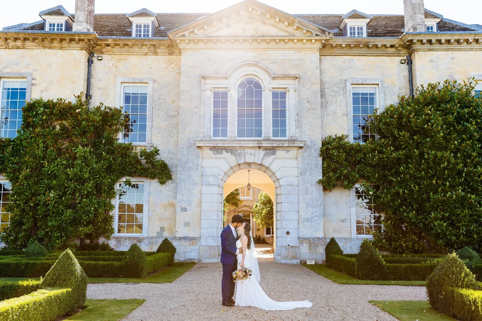A couple embraces in front of a stately manor, framed by lush greenery and manicured gardens, perfect for a romantic outdoor wedding.