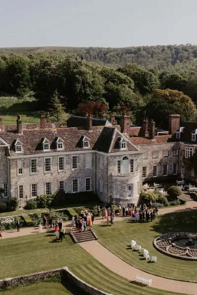 Lush green grounds surround a grand estate, where guests gather for a stylish outdoor ceremony on a sunny day.