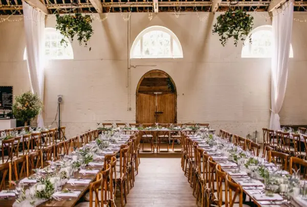 Rustic wedding venue featuring long wooden tables adorned with greenery, elegant decor, and natural light streaming through arched windows.