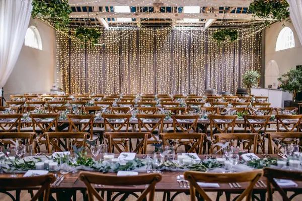 Rustic-chic banquet hall featuring wooden tables, crossback chairs, and twinkling fairy lights draping from the ceiling. Elegant greenery accents enhance the ambiance.