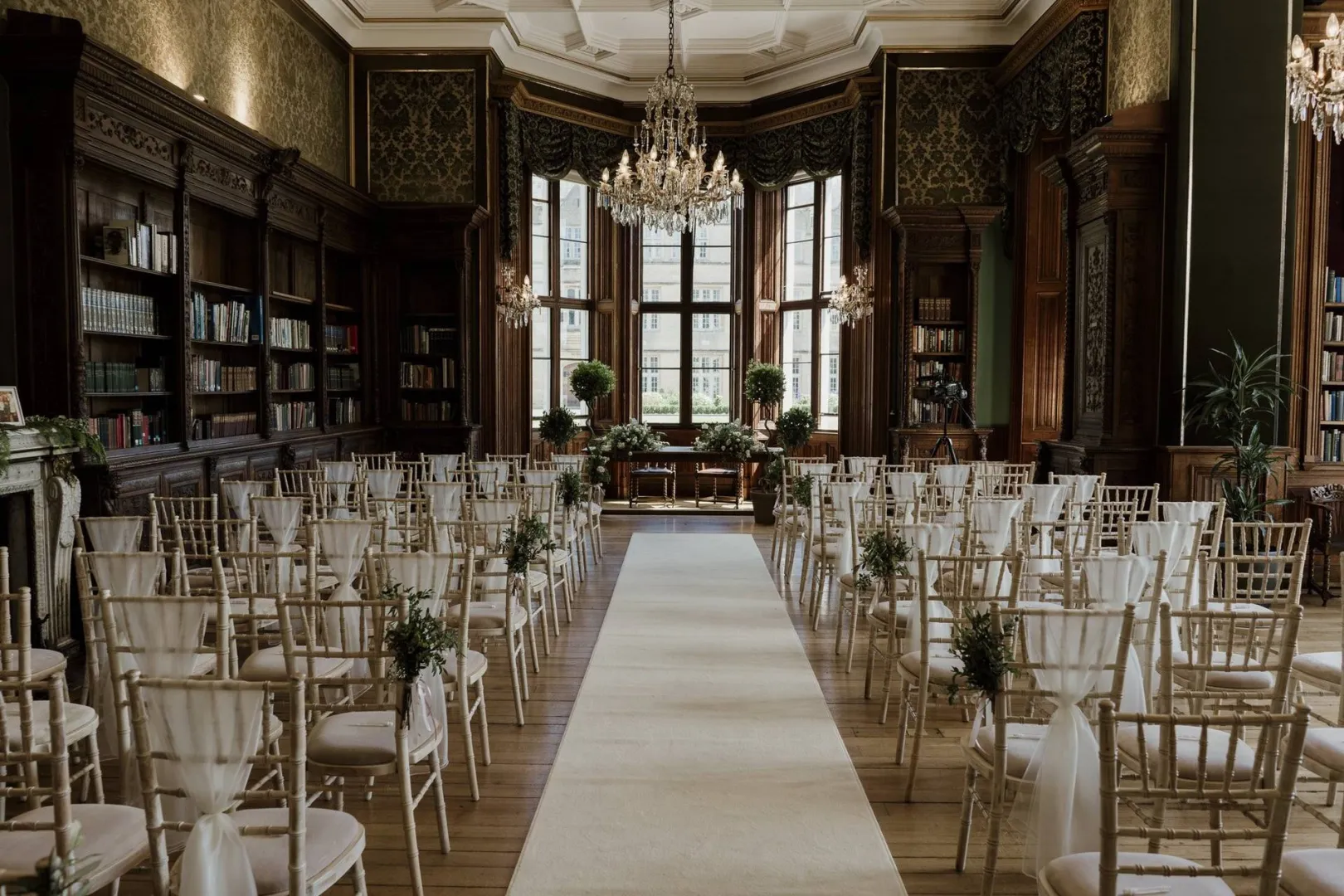 Elegant indoor ceremony space featuring gold Chiavari chairs, a lush carpet aisle, and a backdrop of vintage bookshelves and chandeliers.