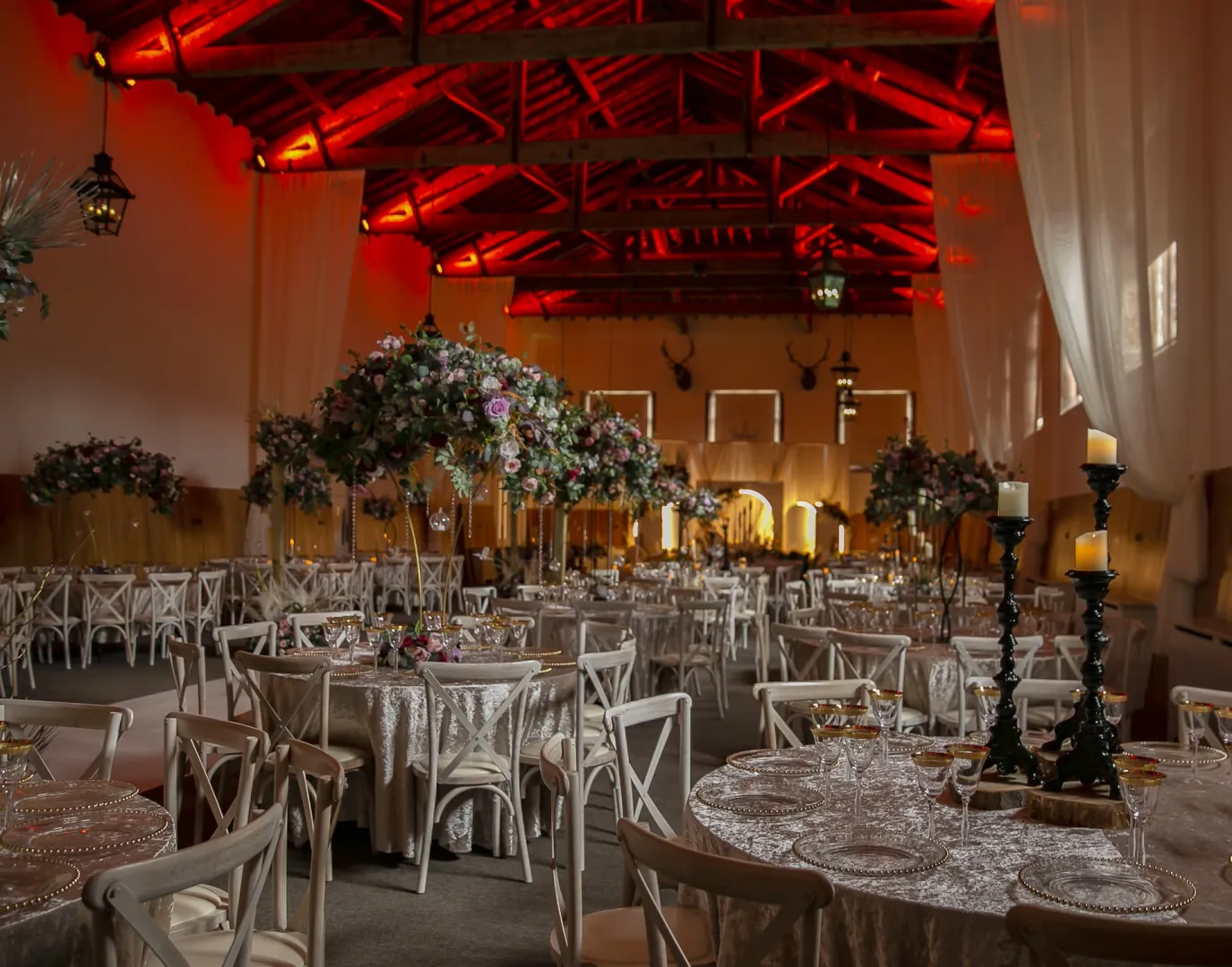 Elegant reception space featuring rustic wooden beams, adorned tables with floral centerpieces, and ambient red lighting for a romantic atmosphere.