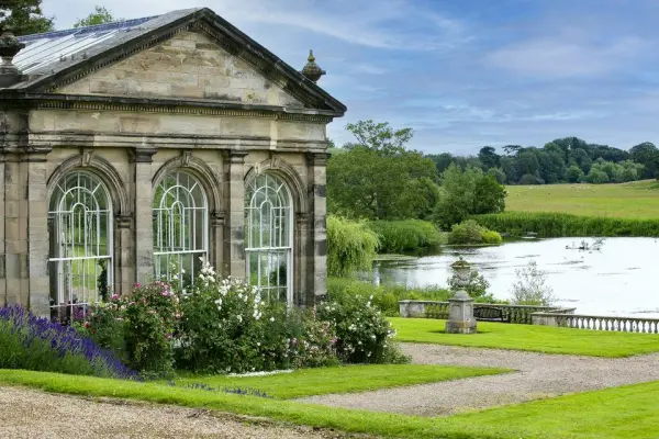 A charming glass-fronted building surrounded by lush gardens, overlooking a serene lake and rolling green fields, perfect for outdoor ceremonies.