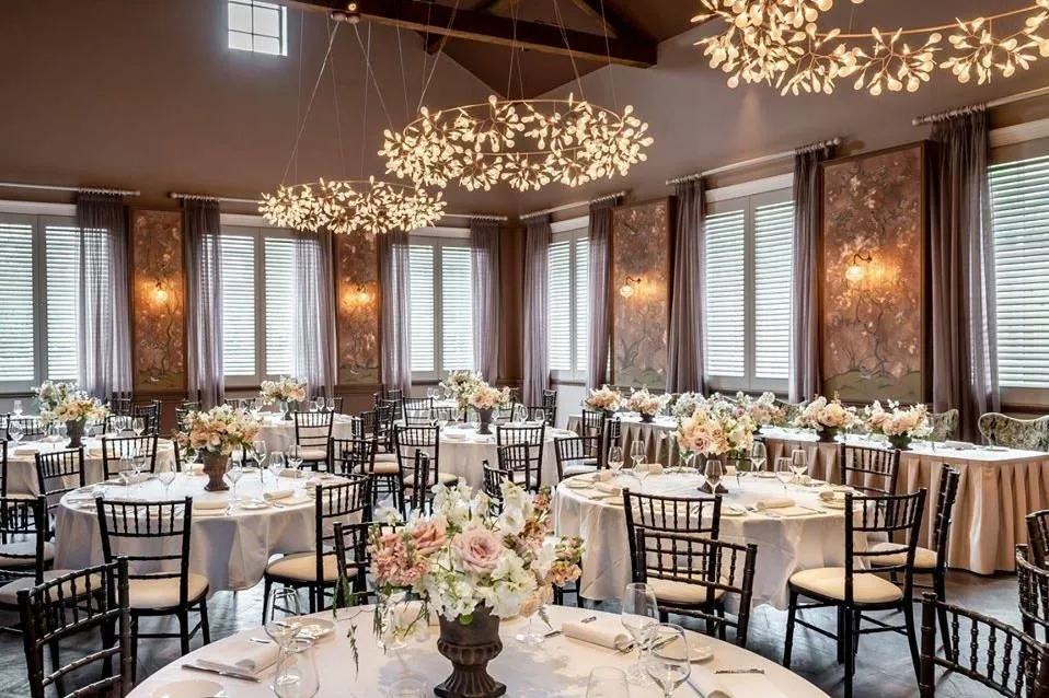 Elegant ballroom featuring round tables adorned with floral centerpieces, chic black chairs, and unique chandelier lighting. Natural light filters through large windows.