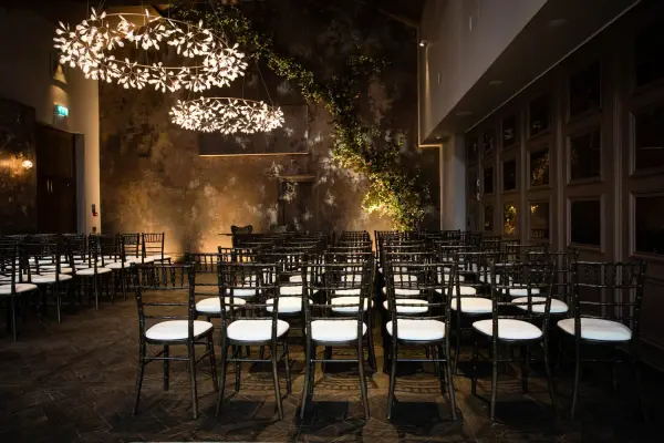 Elegant indoor ceremony space with stylish chandeliers, black chairs, and a textured wall adorned with greenery. Perfect for intimate weddings.