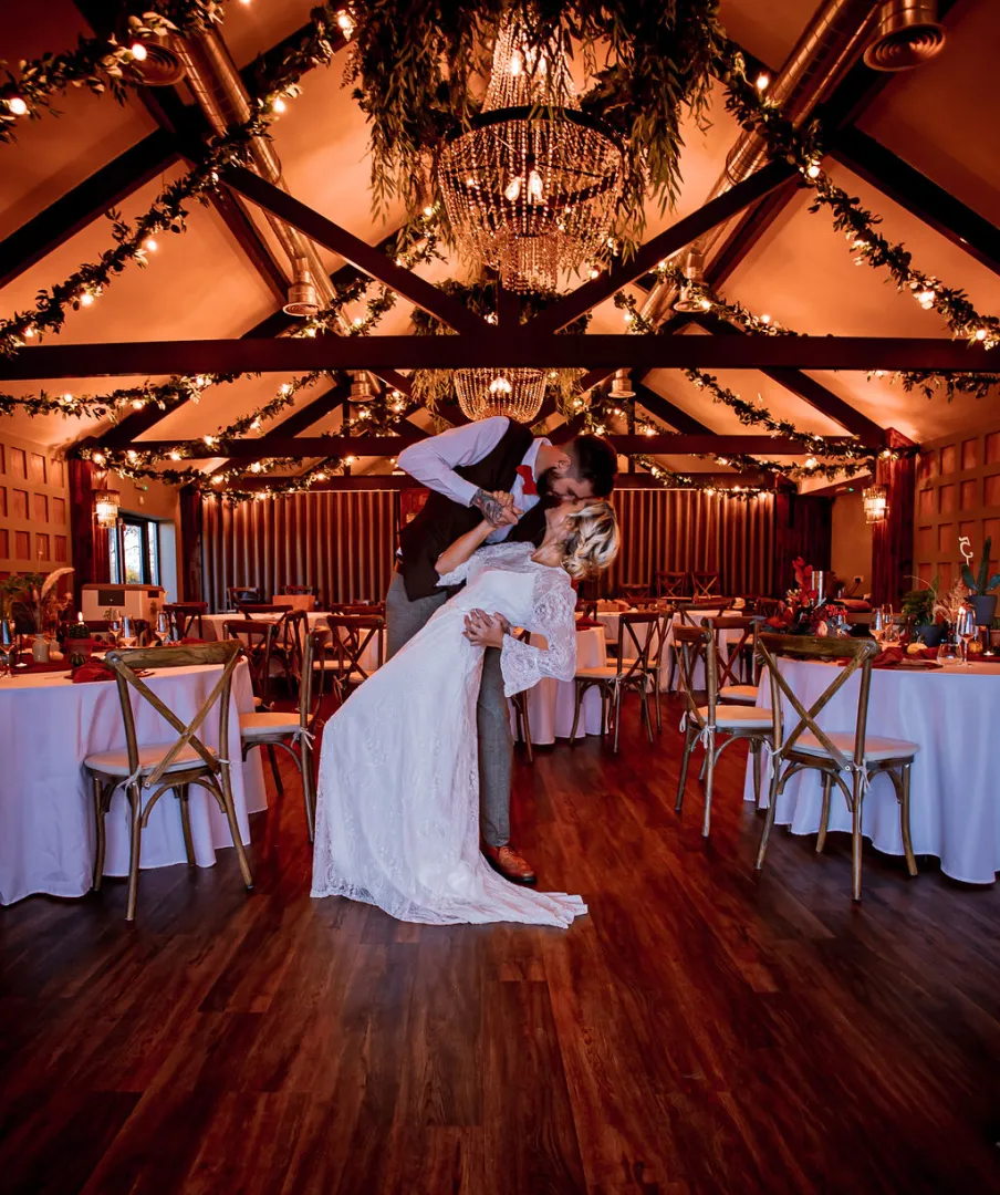 A couple shares a romantic moment in an elegant rustic reception hall, adorned with string lights and floral decor, perfect for celebrations.