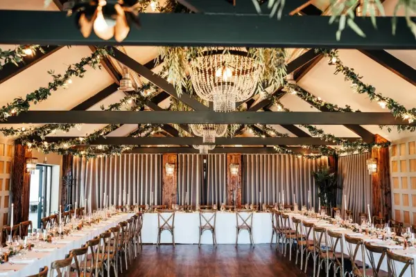 Charming reception space featuring rustic beams, elegant chandeliers, and long dining tables adorned with lush greenery and warm lighting.