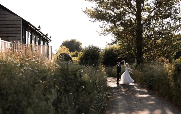 A couple embraces on a scenic pathway lined with wildflowers, leading to a charming rustic venue surrounded by lush greenery.