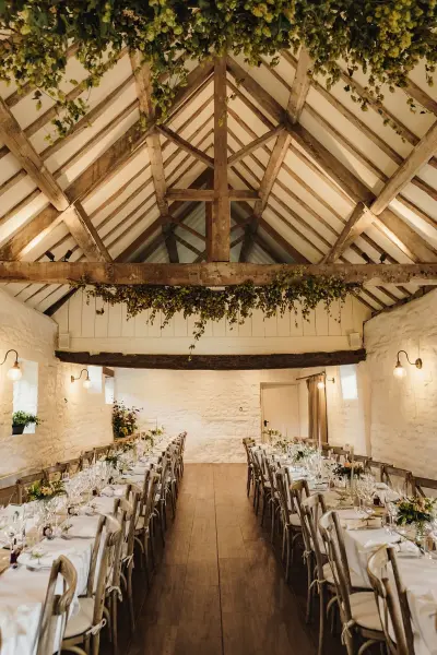 Charming rustic barn interior featuring exposed beams, long dining tables adorned with greenery, and warm ambient lighting. Perfect for intimate receptions.
