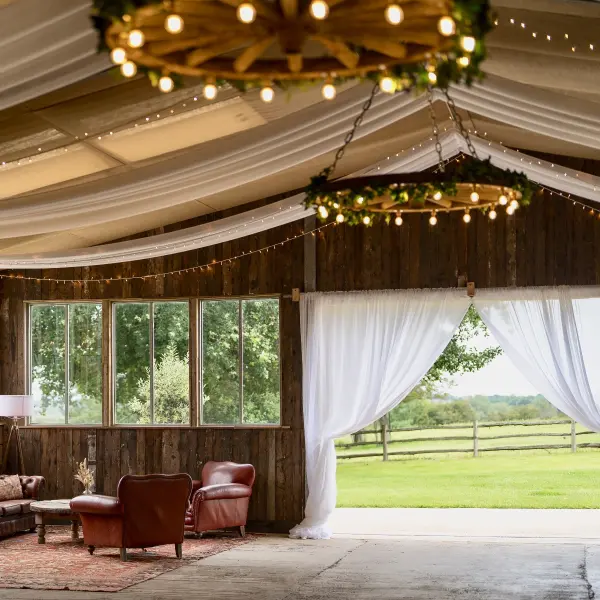 Cozy rustic barn interior featuring elegant draped ceilings, warm string lights, and inviting leather seating with scenic views outside.
