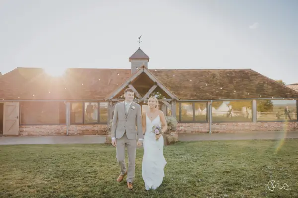 A couple walks hand in hand on a grassy area, with a charming rustic barn featuring large windows and warm sunset light in the background.