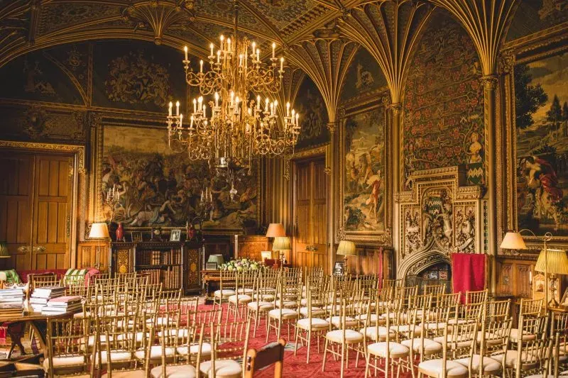 Luxurious ceremony space featuring ornate chandeliers, gilded details, and elegant chairs arranged on plush red carpeting.
