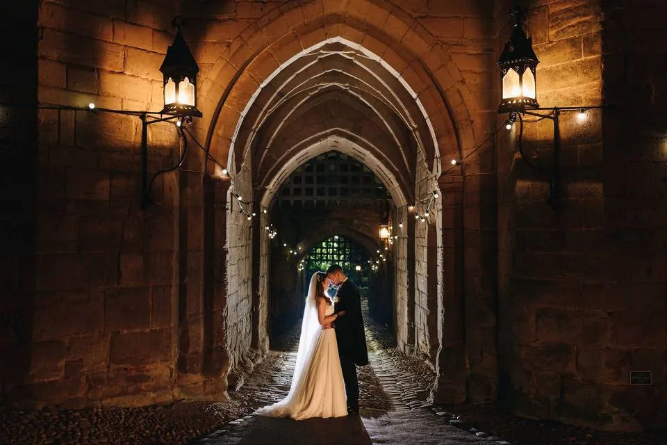 A bride and groom embrace under softly lit arches, with warm lanterns and romantic string lights enhancing the venue's enchanting atmosphere.