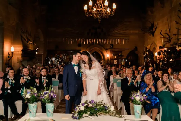 A couple shares a joyful kiss in an ornate venue with elegant decor, surrounded by a cheering audience and vibrant floral arrangements.
