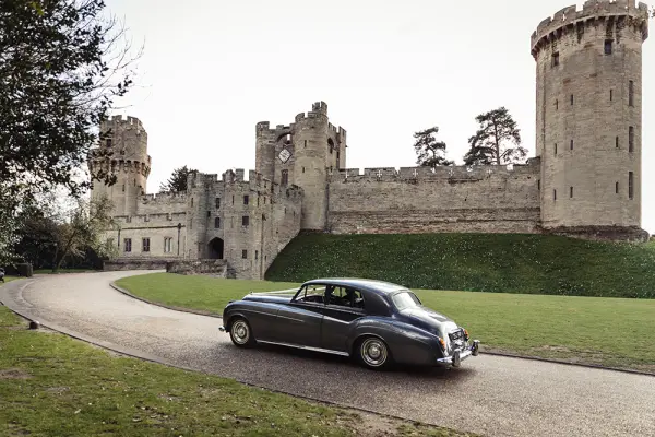 Classic car approaches a stunning stone castle, featuring towering turrets and lush green grounds, perfect for a fairytale wedding backdrop.