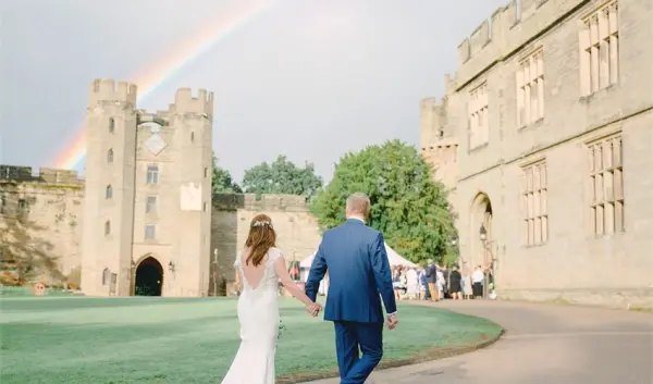 A couple walks hand in hand towards a historic castle, framed by a vibrant rainbow and lush green grounds for a romantic wedding backdrop.