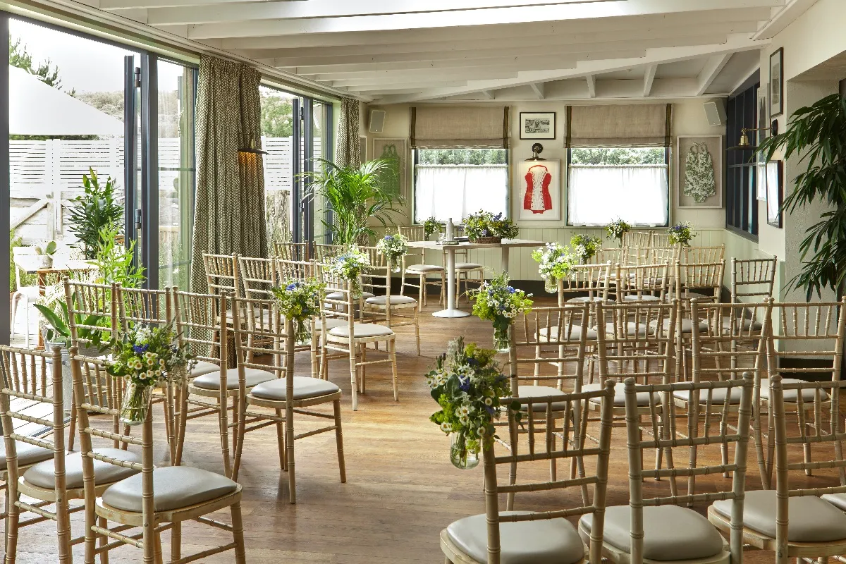 Sunlit indoor ceremony space with elegant wooden chairs, greenery accents, and large windows offering a charming ambiance for weddings.