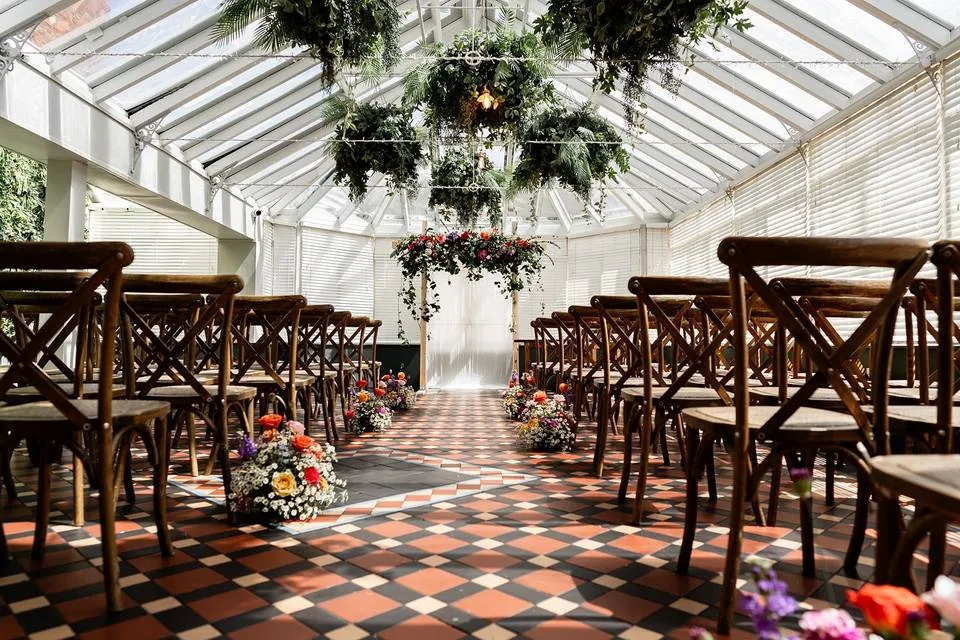 Lush greenery and floral arrangements enhance a charming glass-enclosed ceremony space, perfect for intimate wedding vows.