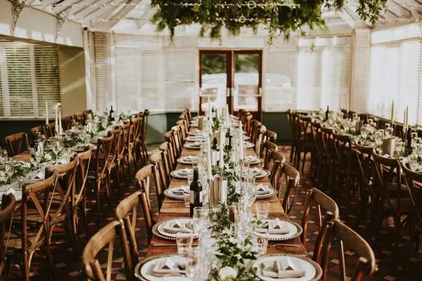 Sunlit reception space featuring long wooden tables decorated with greenery, candles, and elegant place settings for a romantic atmosphere.