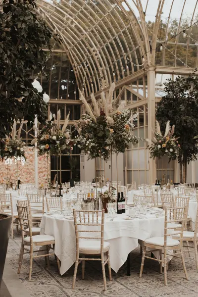 Elegant indoor reception with round tables adorned with white linens, tall floral centerpieces, and a stunning glass atrium ceiling.
