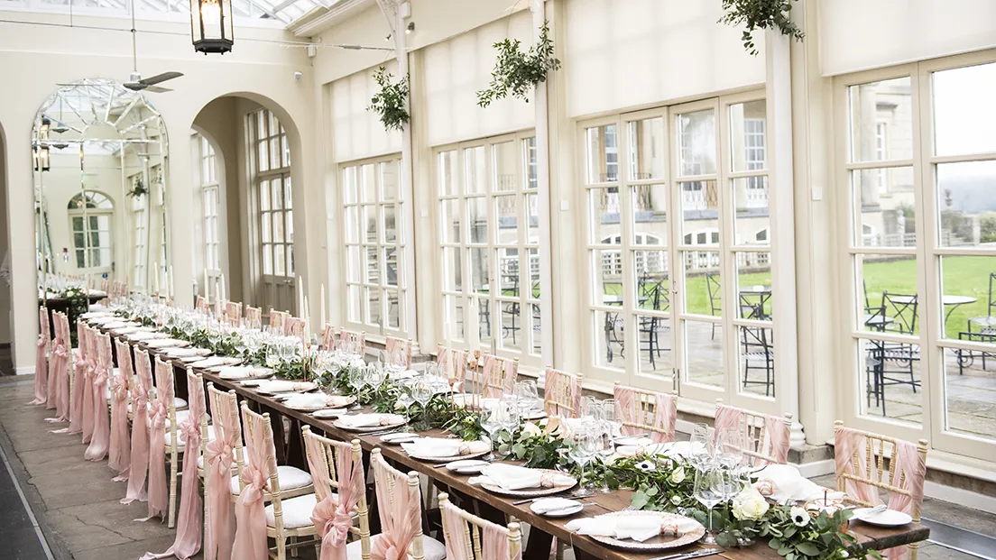 Elegant indoor reception space with long wooden tables, delicate floral centerpieces, and pink satin chair sashes, surrounded by large windows.