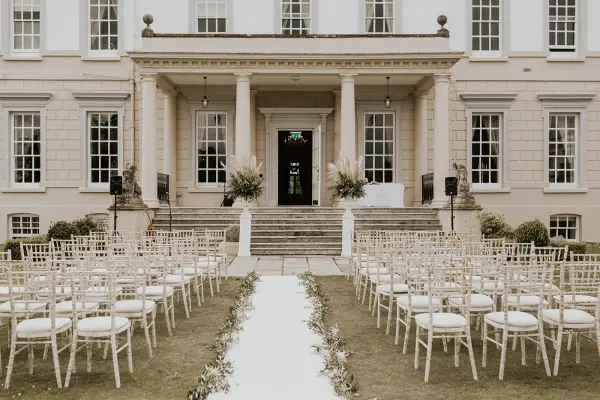 Sunlit outdoor ceremony space with elegant white chairs arranged on a manicured lawn leading to a grand entrance. Ideal for romantic weddings.