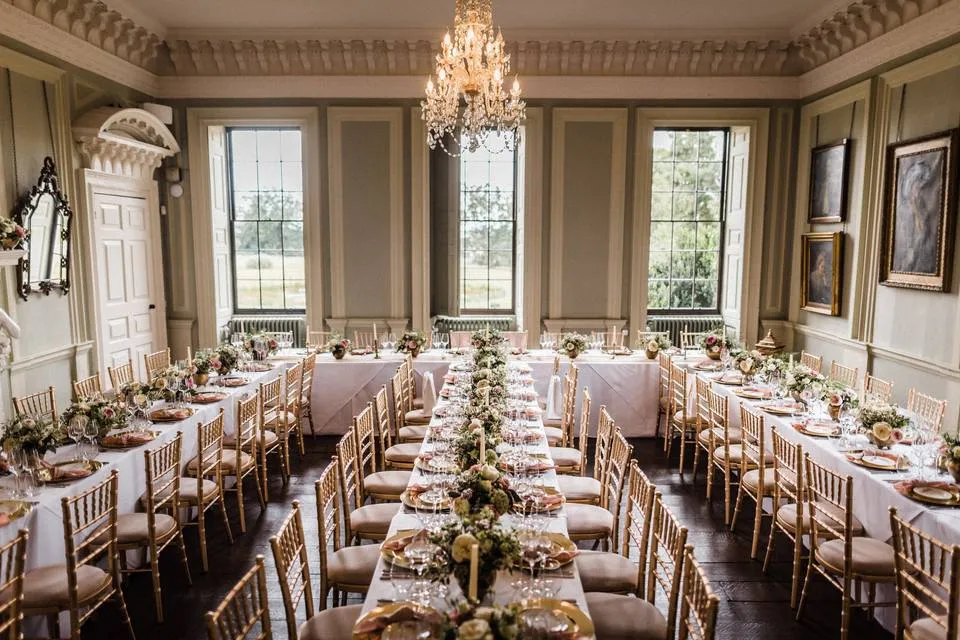 Elegant dining area with long tables set for a wedding, featuring chandeliers, soft lighting, and beautiful floral centerpieces.