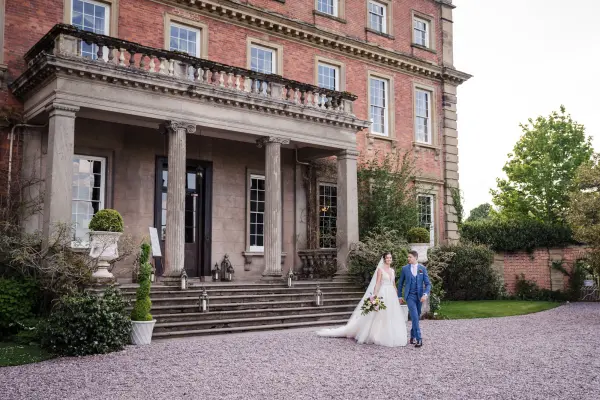 Elegant couple walks hand-in-hand up the grand staircase of a stately mansion, surrounded by manicured gardens and lush greenery.