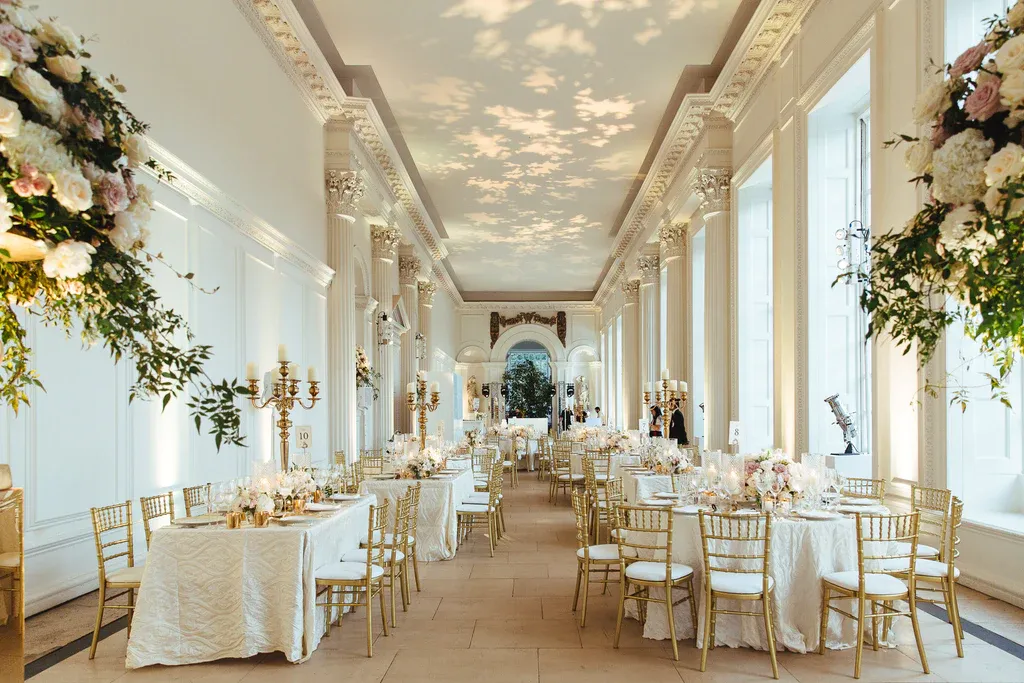 Elegant reception space featuring white walls, tall windows, floral arrangements, and gold chairs, creating a romantic atmosphere for weddings.