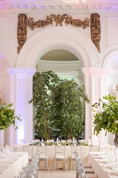 Elegant indoor wedding venue featuring grand columns, lush greenery, and a long dining arrangement under soft lighting.