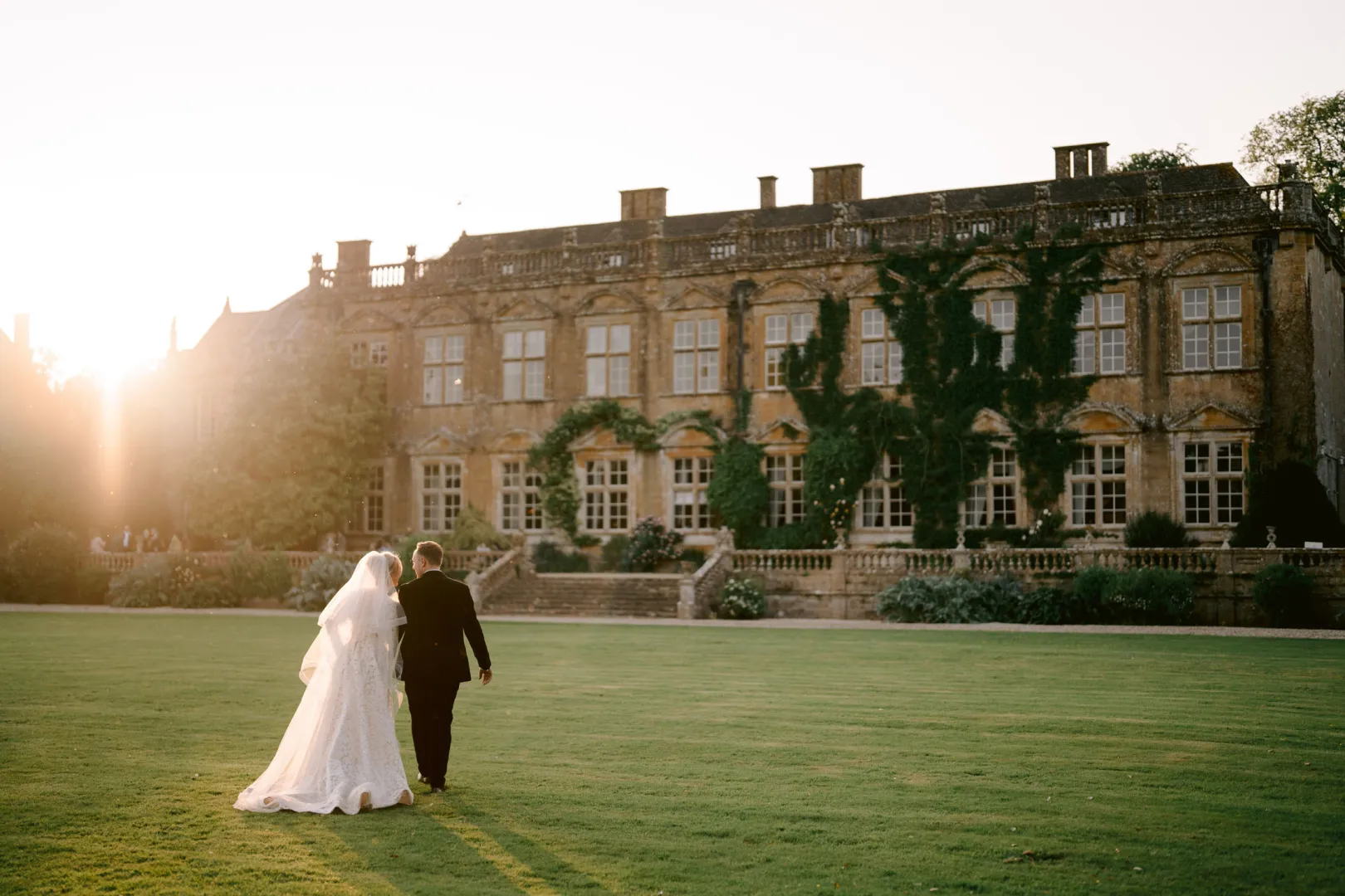 A couple walks hand-in-hand across a lush lawn, with a historic mansion adorned in ivy and glowing in the warm sunset light.