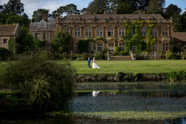 A couple strolls hand-in-hand by a serene pond, with a stunning ivy-covered estate elegantly framing the lush landscape.