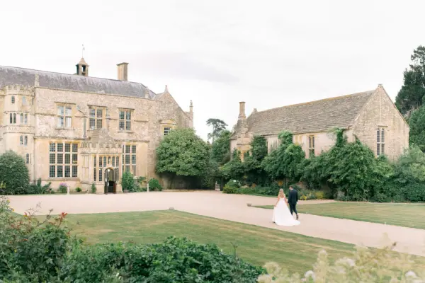 Couple walking hand in hand towards a picturesque stone mansion, surrounded by lush greenery and a beautifully manicured lawn.