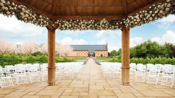 Charming outdoor ceremony space featuring a floral-adorned gazebo, white chairs, and a view of a rustic barn surrounded by lush greenery.