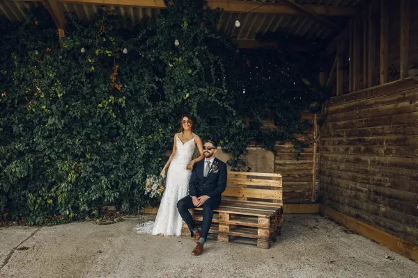 A charming rustic barn backdrop with lush greenery, featuring a bride in a flowing gown and a groom in a suit, seated on a wooden pallet.