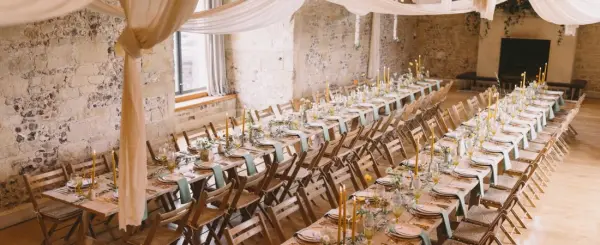 Charming banquet hall featuring rustic stone walls, elegantly set long wooden tables, and soft draped fabric overhead for a romantic atmosphere.