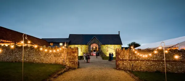 Charming stone entrance leading to a rustic barn venue, adorned with fairy lights and surrounded by lush greenery at twilight.