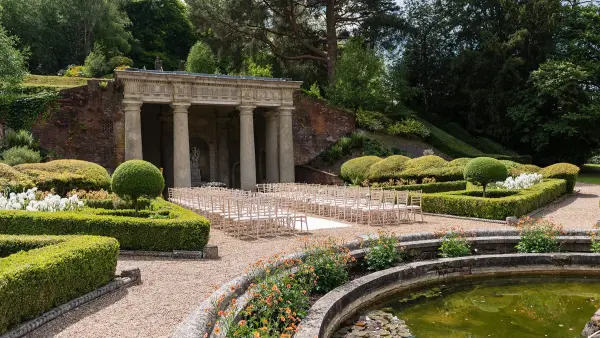Charming outdoor ceremony space with stone columns, manicured hedges, and seating set amidst vibrant gardens and a tranquil pond.