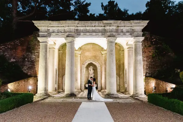 A couple stands together under grand columns at dusk, framed by lush greenery and soft lighting, creating a romantic ceremony backdrop.