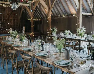 Rustic barn interior with wooden tables set for a wedding reception, adorned with floral centerpieces and twinkling lights overhead.