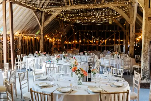Rustic barn interior adorned with fairy lights, elegantly set round tables, and vibrant floral centerpieces, creating a warm reception atmosphere.