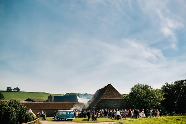 A lively outdoor wedding reception with guests mingling near rustic barns and a vintage blue van under a clear blue sky.