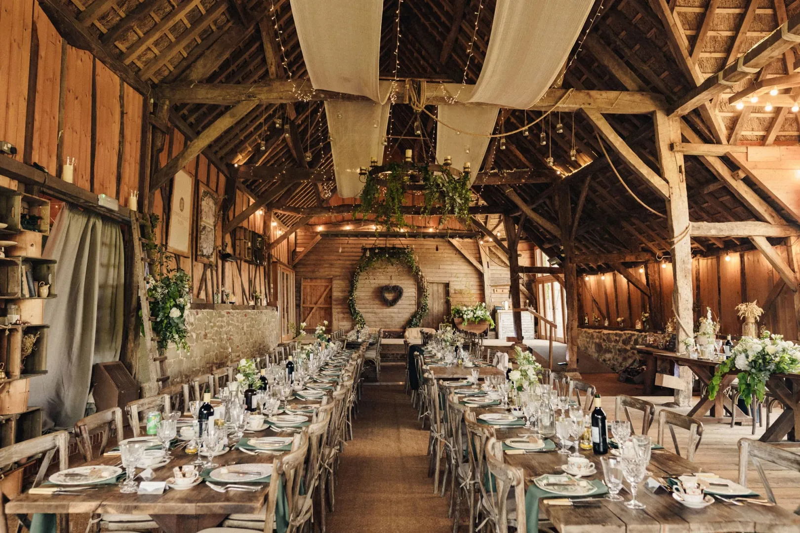 Rustic barn interior adorned with elegant decor, long wooden tables set for a wedding reception with greenery and soft fabric draping overhead.