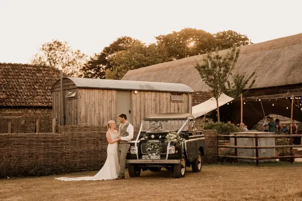 Couple shares a romantic moment by a vintage truck, surrounded by rustic barns and lush greenery, perfect for a charming outdoor wedding.