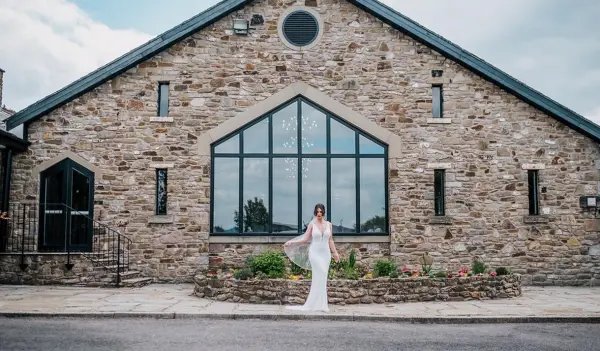 A bride in a sleek wedding gown stands outside a rustic stone venue with large windows and lush flower beds, creating a charming backdrop.