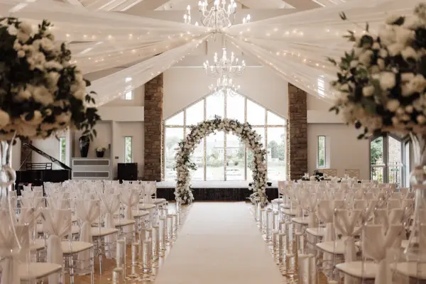 Elegant indoor ceremony space featuring clear chairs, a floral arch, sparkling chandeliers, and beautiful draped ceiling accents.