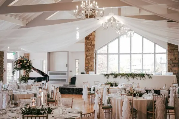 Elegant reception hall with draped ceilings, crystal chandeliers, and beautifully set tables, featuring a grand piano and large windows.